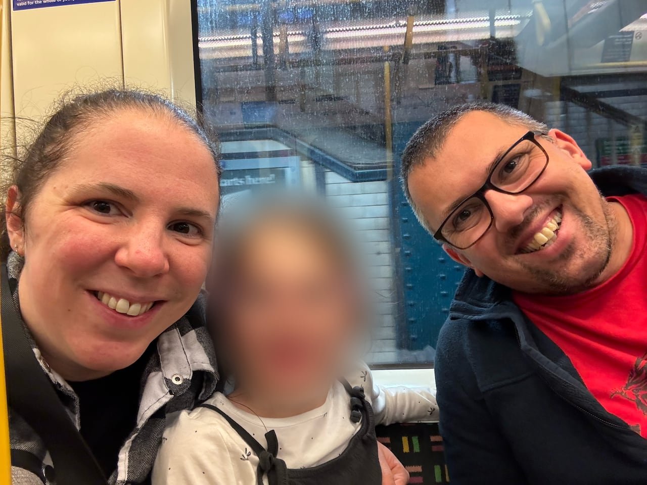 A smiling man, woman and a girl pose for a selfie at a train station. The girl’s face has been blurred.