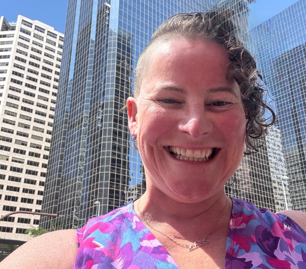 default-419 A woman with short curly hair shaved on one side smiles broadly with tall office buildings in the background.